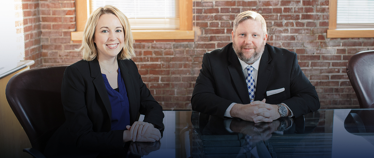 A photograph capturing the interior of the offices, featuring Douglas Ponder and Jaclyn Zimmermann seated and leaning against a glass table. In the background, a brick decorated wall and a window are visible.